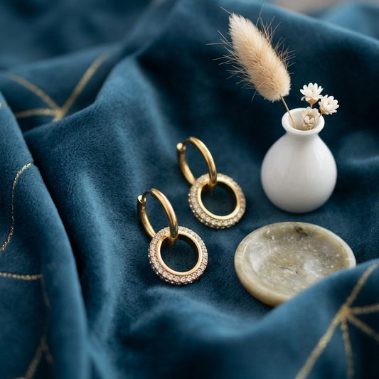 Gold hoop earrings on a blue fabric background with a small white vase and dried flowers.