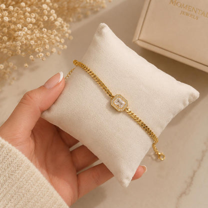 Gold bracelet on a white cushion with a hand holding it, against a neutral background.