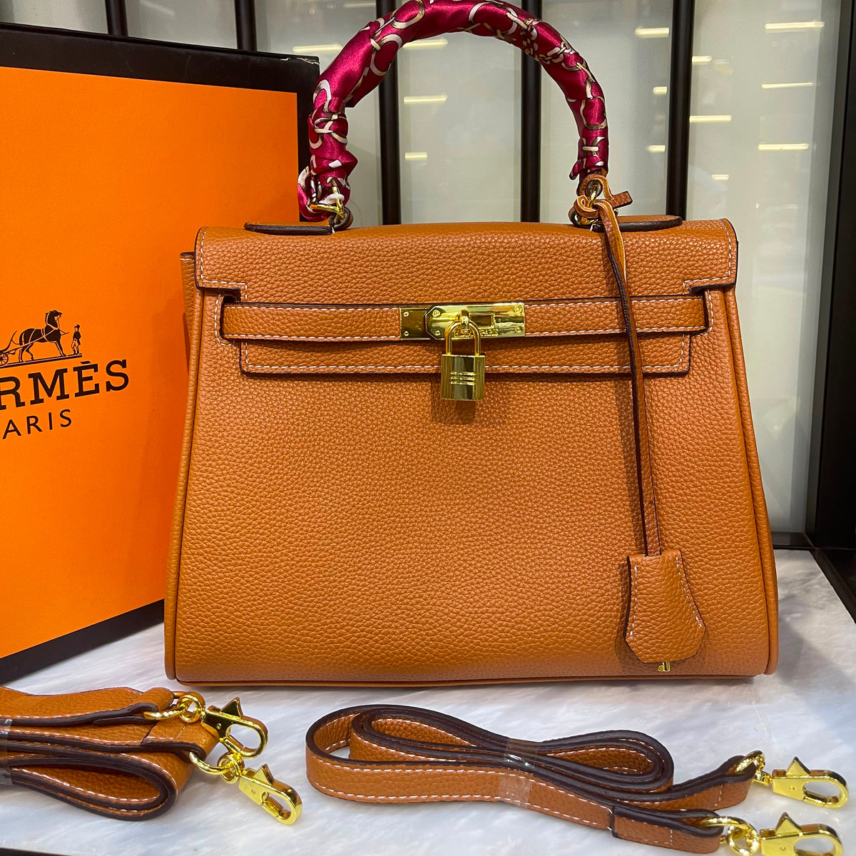 An orange pebbled leather handbag with gold hardware, a red patterned top handle wrap, and an attached leather strap, displayed with an orange Hermès box and a second detachable strap on a marble surface.