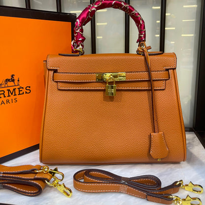 An orange pebbled leather handbag with gold hardware, a red patterned top handle wrap, and an attached leather strap, displayed with an orange Hermès box and a second detachable strap on a marble surface.