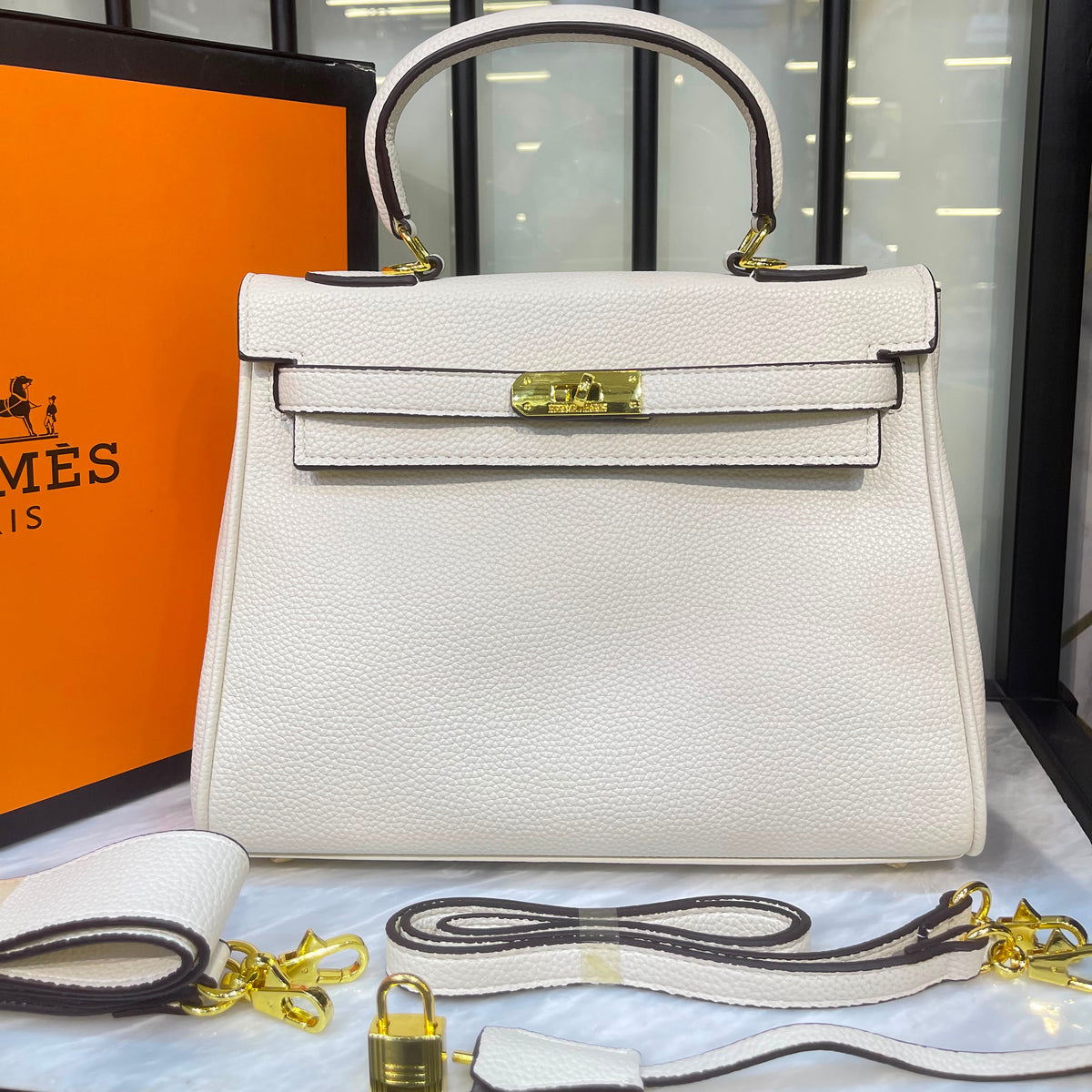 An off-white pebbled leather top-handle handbag with gold-tone hardware, displayed on a marble surface with its strap, lock, and an orange Hermes box in the background.