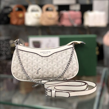 A Goyard Paris shoulder bag in white coated canvas with their signature embossed chevron pattern, featuring a silver-tone chain strap and a matching white leather strap laid out in front. The bag is displayed on a glass shelf with a blurred background of other handbags.
