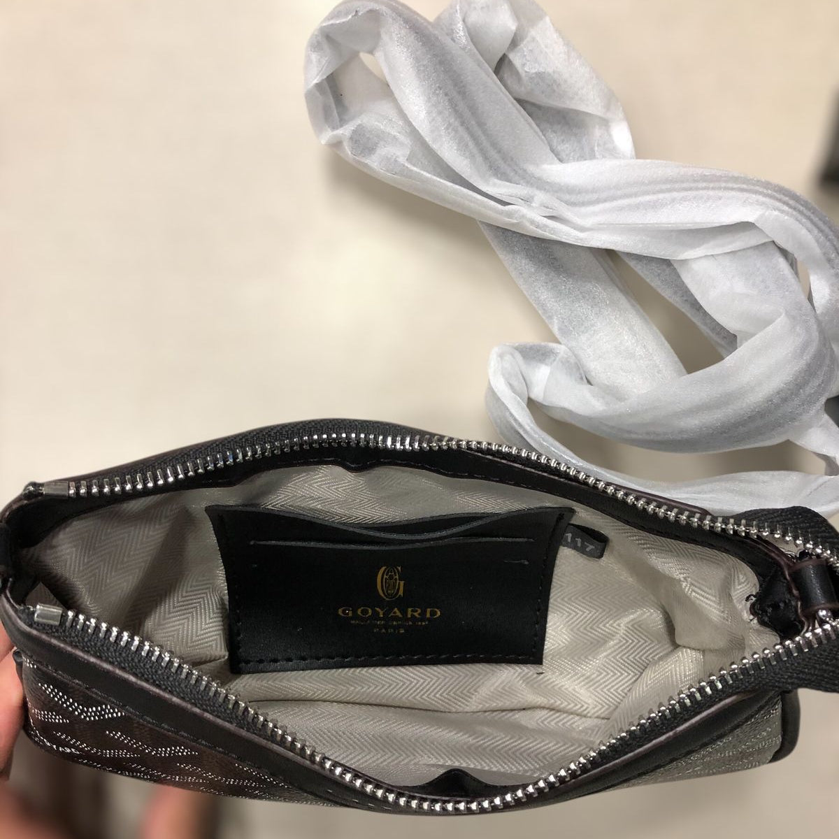 An open black quilted Goyard shoulder bag with a silver zipper, showing a light gray interior lining and a black brand label. A white fabric strap is draped above the bag, and a hand is holding it at the bottom left.